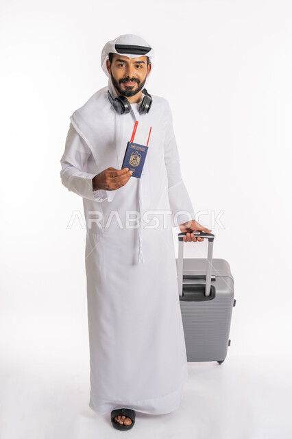 إيماءات السرور والسعادة، القيام بالجولات الترفيهية، مفهوم السياحة في إجازة الصيف، بورتريه لرجل عربي خليجي اماراتي يرتدي الكندورة والغترة وسماعة الرأس يحمل بيده جواز السفر وتيكت الطيارة يمسك بحقيبة الامتعة، صورة لكامل الجسم، خلفية بيضاء
