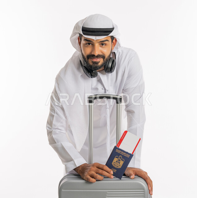 الاستعداد لقضاء رحلة صيفية ممتعة، القيام بالجولات الترفيهية، مفهوم السياحة في إجازة الصيف، بورتريه لرجل عربي خليجي اماراتي يرتدي الكندورة والغترة وسماعة الرأس يحمل بيده جواز السفر وتيكت الطيارة يقوم بالاتكاء على حقيبة الامتعة، خلفية بيضاء