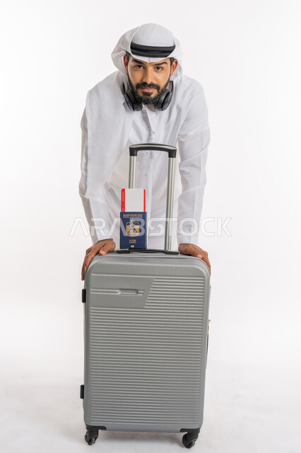 الاستعداد لقضاء رحلة صيفية ممتعة، القيام بالجولات الترفيهية، مفهوم السياحة في إجازة الصيف، بورتريه لرجل عربي خليجي اماراتي يرتدي الكندورة والغترة وسماعة الرأس يحمل بيده جواز السفر وتيكت الطيارة يقوم بالاتكاء على حقيبة الامتعة، خلفية بيضاء
