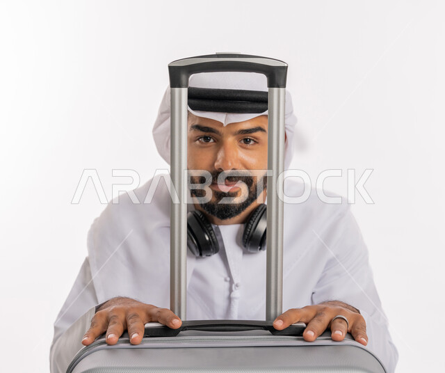 ايماءات السرور والسعادة، الاستعداد لقضاء رحلة صيفية ممتعة، القيام بالجولات الترفيهية، مفهوم السياحة في إجازة الصيف، بورتريه لرجل عربي خليجي اماراتي يرتدي الكندورة والغترة وسماعة الرأس يقوم بالاختباء خلف حقيبة الامتعة، خلفية بيضاء