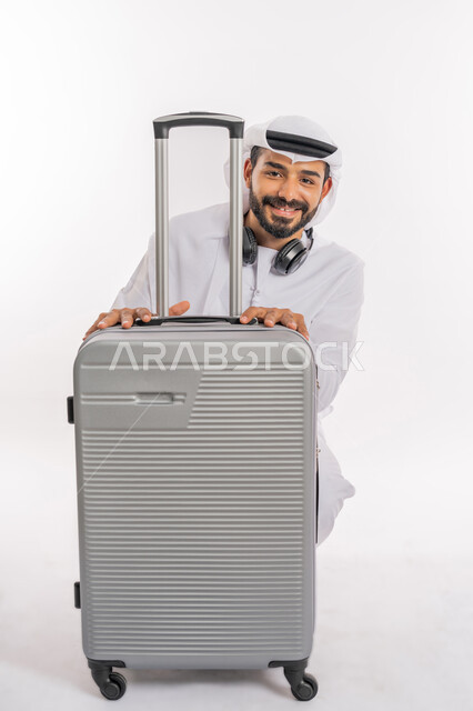 تعابير السرور والسعادة، الاستعداد لقضاء رحلة صيفية ممتعة، القيام بالجولات الترفيهية، مفهوم السياحة في إجازة الصيف، بورتريه لرجل عربي خليجي اماراتي يرتدي الكندورة والغترة وسماعة الرأس يقوم بالاختباء خلف حقيبة الامتعة، خلفية بيضاء