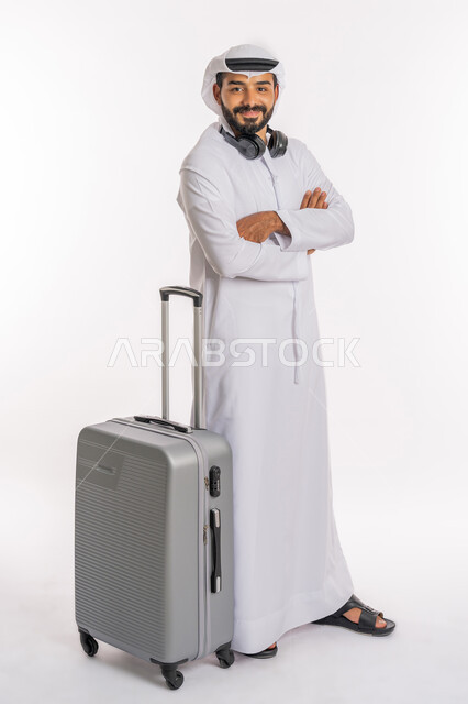 ايماءات الثقة بالنفس، الاستعداد لقضاء رحلة صيفية ممتعة، القيام بالجولات الترفيهية، مفهوم السياحة في إجازة الصيف، بورتريه لرجل عربي خليجي اماراتي يرتدي الكندورة والغترة وسماعة الرأس يقف بايدي متقاطعة بجانب حقيبة الامتعة، صورة لكامل الجسم، خلفية بيضاء