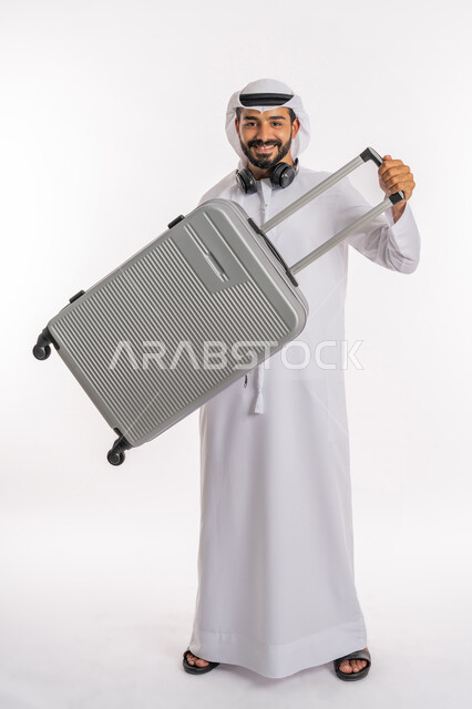 القيام بالجولات الترفيهية، النظر إلى الكاميرا بإيماءات السرور والسعادة، الاستعداد لقضاء رحلة صيفية ممتعة، مفهوم السياحة في إجازة الصيف، بورتريه لرجل عربي خليجي اماراتي يرتدي الكندورة والغترة وسماعة الرأس يحمل بيده حقيبة الامتعة، صورة لكامل الجسم، خلفية بيضاء