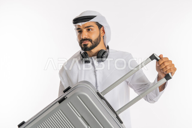 مفهوم السياحة في إجازة الصيف، القيام بالجولات الترفيهية، إيماءات السرور والسعادة، الاستعداد لقضاء رحلة صيفية ممتعة، بورتريه مقرب لرجل عربي خليجي اماراتي يرتدي الكندورة والغترة وسماعة الرأس يحمل بيده حقيبة الامتعة، خلفية بيضاء