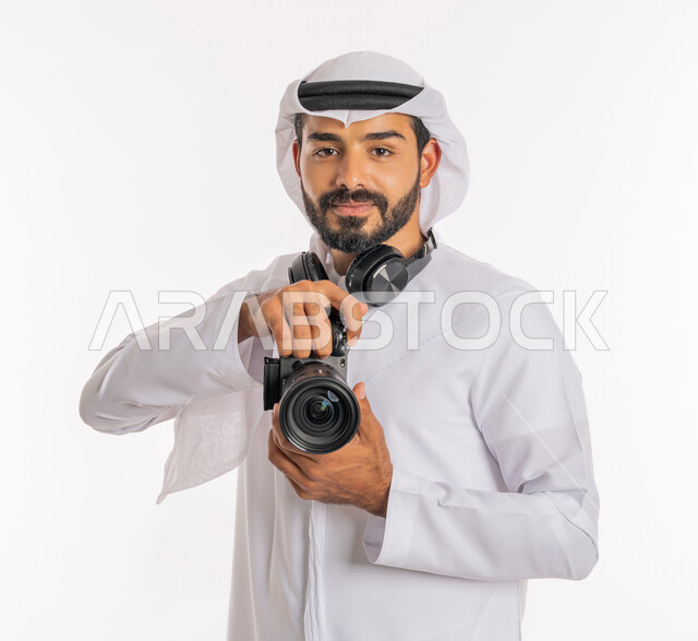 التقط الصور التذكارية، فعاليات وأنشطة ترفيهية سياحية، كاميرات حديثة ومتطورة، استخدام التقنيات التكنولوجية في مجال التصوير، بورتريه مقرب لرجل عربي خليجي اماراتي يرتدي الكندورة والغترة وسماعة الرأس يحمل بيده كاميرا عالية الدقة، خلفية بيضاء