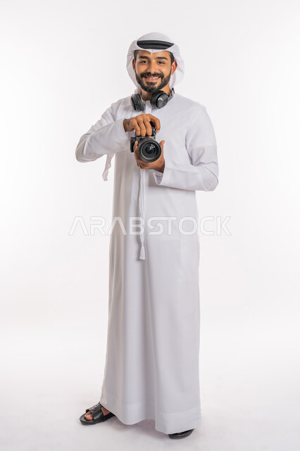 Modern and advanced cameras, tourist entertainment events and activities, take souvenir photos, use of technological techniques in the field of photography, a portrait of an Arab Gulf Emirati man wearing a kandora, a ghutra, and a headset, holding a high-resolution camera in his hand, a full-body photo, a white background.