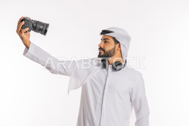 فعاليات وأنشطة ترفيهية سياحية، استخدام التقنيات التكنولوجية في مجال التصوير، كاميرات حديثة ومتطورة، بورتريه مقرب لرجل عربي خليجي اماراتي يرتدي الكندورة والغترة وسماعة الرأس يحمل بيده كاميرا عالية الدقة يقوم بالتقاط صورة سيلفي تذكارية، خلفية بيضاء