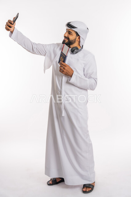 إيماءات الفرح والسعادة، مفهوم السياحة والسفر، القيام بالجولات الترفيهية في وقت الإجازة، بورتريه لرجل عربي خليجي اماراتي يرتدي الكندورة والغترة والسماعات يحمل بيده الباسبور وتيكت الطيارة يقوم بالتقاط صورة سيلفي، صورة لكامل الجسم، خلفية بيضاء