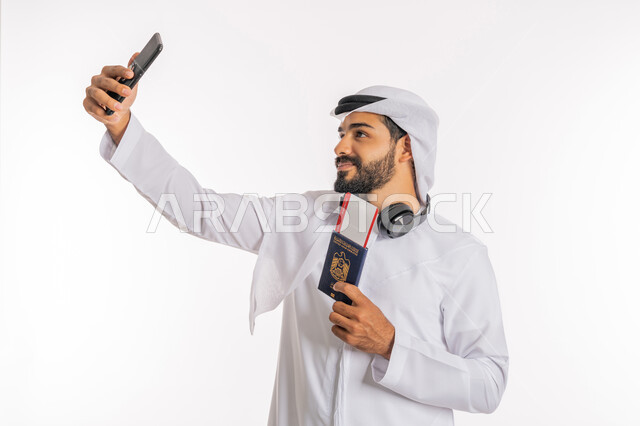 تعابير السرور والسعادة، مفهوم السياحة والسفر، القيام بالجولات الترفيهية في وقت الإجازة، بورتريه مقرب لرجل عربي خليجي اماراتي يرتدي الكندورة والغترة والسماعات يحمل بيده الباسبور وتيكت الطيارة يقوم بالتقاط صورة سيلفي، خلفية بيضاء