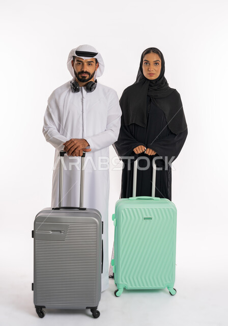 انتظار موعد الطائرة، الاستعداد لقضاء رحلة صيفية ممتعة، مفهوم السياحة والقيام بالجولات الترفيهية في وقت الإجازة، بورتريه لرجل عربي خليجي اماراتي يرتدي الكندورة والغترة يمسك بحقيبة الامتعة يقف برفقة زوجته، صورة الجسم بالطول الكامل، خلفية بيضاء