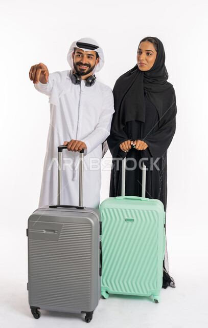 الاستعداد لقضاء رحلة صيفية ممتعة، مفهوم السياحة والقيام بالجولات الترفيهية في وقت الإجازة، انتظار موعد الطائرة، بورتريه لرجل عربي خليجي اماراتي يرتدي الكندورة والغترة يمسك بحقيبة الامتعة يقف برفقة زوجته يشير باصبعه لمكان ما، صورة الجسم بالطول الكامل، خلفية بيضاء