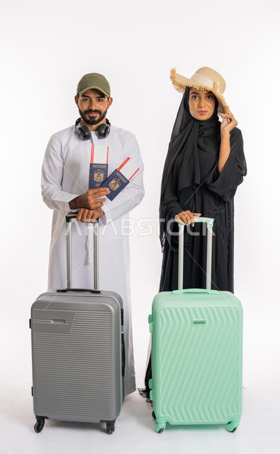 الاستعداد لقضاء رحلة صيفية ممتعة، انتظار موعد الطائرة، بورتريه لزوجان عربيان اماراتيان يرتديان القبعات يحملان جواز وتأشيرة السفر يمسكان بحقائب الامتعة، مفهوم السياحة والقيام بالجولات الترفيهية في وقت الإجازة، صورة لكامل الجسم، خلفية بيضاء