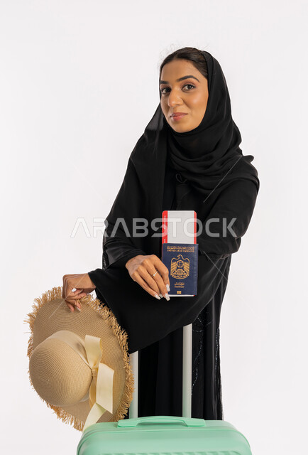 الاستعداد لقضاء رحلة صيفية ممتعة، مفهوم السياحة والقيام بالجولات الترفيهية في وقت الإجازة، شابة سعودية أنيقة، بورتريه لسيدة عربية خليجية اماراتية محجبة ترتدي عباءة سوداء تحمل جواز وتأشيرة السفر تمسك بيدها حقيبة الامتعة وقبعة قشية، خلفية بيضاء