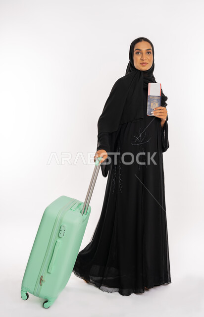 القيام بالجولات الترفيهية في وقت الإجازة، الاستعداد لقضاء رحلة صيفية ممتعة، مفهوم السياحة، شابة سعودية أنيقة، بورتريه لسيدة عربية خليجية اماراتية محجبة ترتدي عباءة سوداء تحمل جواز وتأشيرة السفر تمسك بيدها حقيبة سفر، خلفية بيضاء