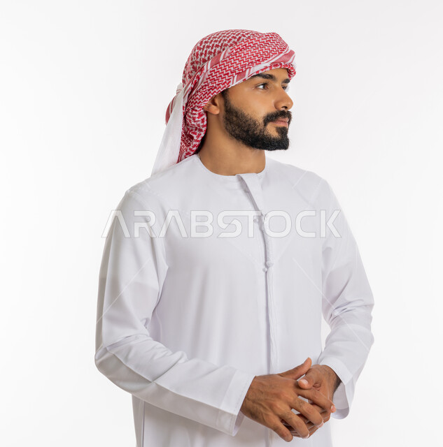 مفهوم الأناقة والرجولة، الاهتمام بالمظهر الخارجي، بورتريه مقرب لشاب عربي خليجي اماراتي أنيق يرتدي الكندورة والعمامة يقف بايدي متشابكة، النظر إلى مكان ما بإيماءات التركيز، خلفية بيضاء