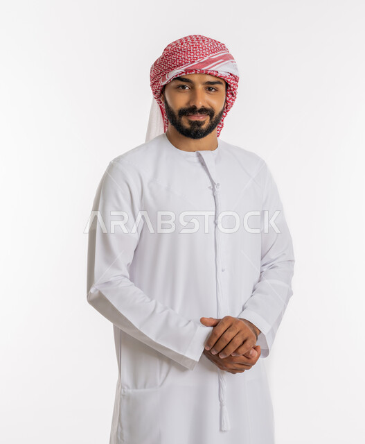 تعابير السرور والفرح، الأناقة والاهتمام بالمظهر الخارجي، بورتريه لشاب عربي خليجي اماراتي أنيق يرتدي الكندورة والعمامة يقف بايدي متشابكة، النظر إلى الكاميرا بإيماءات السعادة، مفهوم الثقة بالنفس، خلفية بيضاء