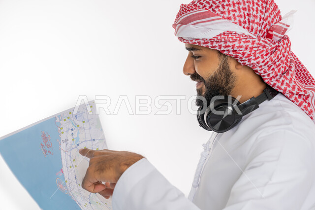 تحديد مسار سير الرحلة، بورتريه مقرب من الجانب لشاب عربي خليجي سعودي يرتدي الكندورة والعمامة والسماعات يقوم بالاشارة باصبعه لمكان ما على الخريطة بإيماءات وجه تدل على السعادة، السياحة والسفر ورحلات السفاري، مفهوم البحث والاستكشاف حول العالم، خلفية بيضاء