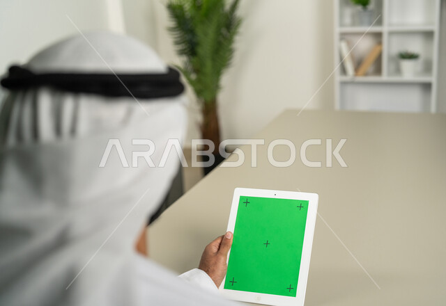 Communicating with family and relatives via tablet applications, displaying a blank green chroma screen on technical devices, using modern and advanced technology for learning, a picture from behind of an Arab Gulf Emirati man wearing a kandora and ghutra holding a tablet in his hand browsing social media.