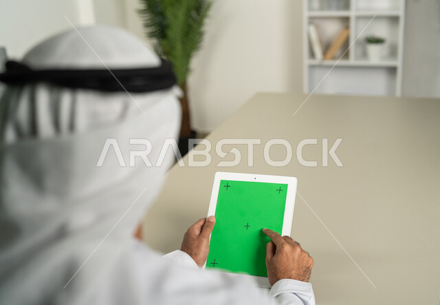 الدردشات والمحادثات مع العائلة والأصدقاء, عرض شاشة  كروما خضراء فارغة على الأجهزة التقنية، استعمال التكنولوجيا الحديثة والمتطورة للتعلم، صورة من الخلف لرجل عربي خليجي اماراتي يرتدي الكندورة  والغترة يمسك بيده تابلت يقوم بتصفح وسائل التواصل الإجتماعي
