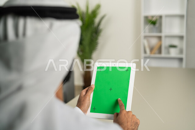 مشاهدة البرامج المفضلة في اوقات الفراغ, عرض شاشة  كروما خضراء فارغة على الأجهزة التقنية، استعمال التكنولوجيا الحديثة والمتطورة للتعلم، صورة من الخلف لرجل عربي خليجي اماراتي يرتدي الكندورة  والغترة يمسك بيده تابلت يقوم بتصفح وسائل التواصل الإجتماعي