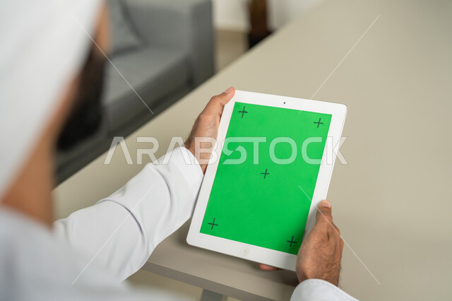 Completing work and tasks, displaying a blank green chroma screen on technical devices, using modern and advanced technology for learning, a picture from behind of an Arab Gulf Emirati man wearing a kandora and ghutra, holding a tablet in his hand, following social media news.