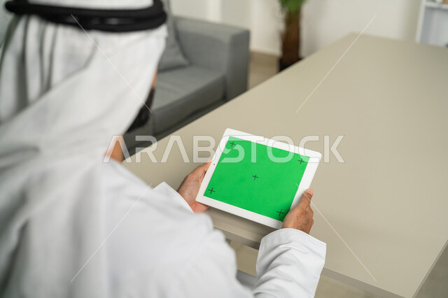 التصفح للصور والفيديوهات بسهولة، عرض شاشة  كروما خضراء فارغة على الأجهزة التقنية، استعمال التكنولوجيا الحديثة والمتطورة للتعلم، صورة من الخلف لرجل عربي خليجي اماراتي يرتدي الكندورة  والغترة يمسك بيده تابلت يقوم بمتابعة أخبار وسائل التواصل الإجتماعي