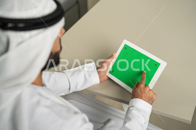 إجراء مكالمة فيديو، عرض شاشة  كروما خضراء فارغة على الأجهزة التقنية، استعمال التكنولوجيا الحديثة والمتطورة للتعلم، صورة من الخلف لرجل عربي خليجي اماراتي يرتدي الكندورة  والغترة يمسك بيده تابلت يقوم بمتابعة أخبار وسائل التواصل الإجتماعي