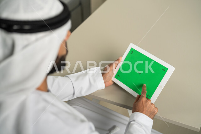 القيام بإجتماعات العمل أونلاين، عرض شاشة  كروما خضراء فارغة على الأجهزة التقنية، استعمال التكنولوجيا الحديثة والمتطورة للتعلم، صورة من الخلف لرجل عربي خليجي اماراتي يرتدي الكندورة  والغترة يمسك بيده تابلت يقوم بمتابعة أخبار وسائل التواصل الإجتماعي