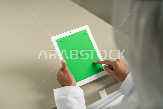 القيام بمقابلات وإجتماعات العمل أونلاين، عرض شاشة  كروما خضراء فارغة على الأجهزة التقنية، استعمال التكنولوجيا الحديثة والمتطورة للتعلم، صورة من الخلف لرجل عربي خليجي اماراتي يرتدي الكندورة  والغترة يمسك بيده تابلت يقوم بمتابعة أخبار وسائل التواصل الإجتماعي