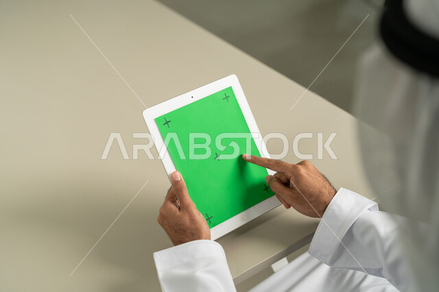 استعمال الجهاز اللوحي في إنجاز العمل، عرض شاشة  كروما خضراء فارغة على الأجهزة التقنية، استعمال التكنولوجيا الحديثة والمتطورة للتعلم، صورة من الخلف لرجل عربي خليجي اماراتي يرتدي الكندورة  والغترة يمسك بيده تابلت يقوم بمتابعة أخبار وسائل التواصل الإجتماعي