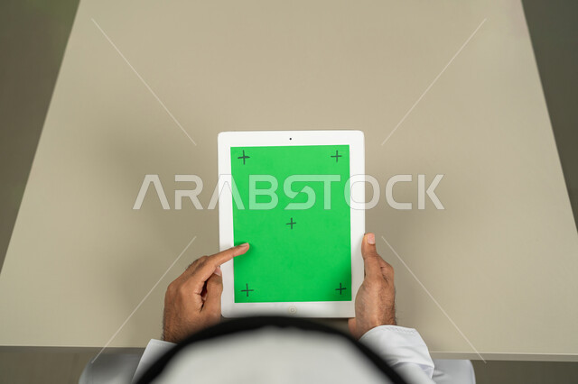 إجراء اجتماعات العمل أونلاين، عرض شاشة  كروما خضراء فارغة على الأجهزة التقنية، استعمال التكنولوجيا الحديثة والمتطورة للتعلم عن بعد، صورة من الاعلى لرجل عربي خليجي اماراتي يرتدي الكندورة  والغترة يمسك بيده تابلت يقوم بمتابعة أخبار وسائل التواصل الإجتماعي