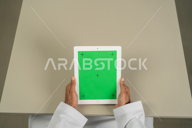 إجراء مقابلات وإجتماعات العمل أونلاين، عرض شاشة  كروما خضراء فارغة على الأجهزة التقنية، استعمال التكنولوجيا الحديثة والمتطورة للتعلم عن بعد، صورة من الاعلى لرجل عربي خليجي اماراتي يمسك بيده تابلت يقوم بمتابعة أخبار وسائل التواصل الإجتماعي