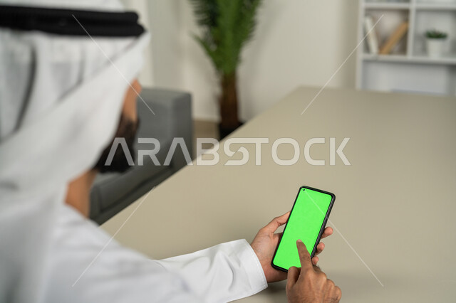 Getting work done quickly and easily, displaying a blank green chroma screen on the mobile phone, integrating technology and technology into practical life, a picture from behind of an Arab Gulf Emirati man holding a mobile phone in his hand, using modern technologies to complete tasks remotely, holding meetings via the Internet