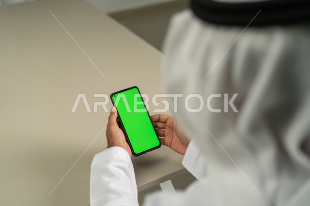 عرض شاشة كروما خضراء فارغة على الهاتف المحمول، دمج التقنية والتكنولوجيا في الحياة العملية، صورة من الخلف لرجل عربي خليجي اماراتي يحمل بيده جوال محمول، استخدام التقنيات الحديثة في انجاز المهام عن بعد، عقد الاجتماعات عن طريق الانترنت
