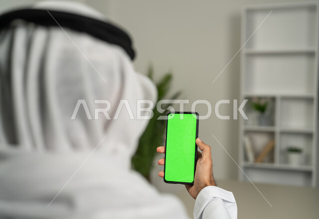 Integrating technology and technology into practical life, browsing social media news, a picture from behind of an Arab Gulf Emirati man using a mobile phone with a green, blank screen, chroma, chatting and conversations with friends, marketing a website