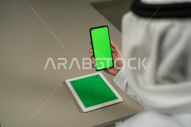 Holding meetings via the Internet, browsing social media news, technical developments and modern technology, a picture from the back of an Arab Gulf Emirati man using a mobile phone and tablet with a blank green screen, chroma, chatting and conversations with friends, marketing a website.