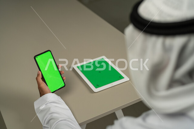 أجهزة الكترونية بشاشة خضراء فارغة كروما، تصفح أخبار وسائل التواصل الإجتماعي، التطورات التقنية والتكنولوجيا الحديثة، صورة من الخلف لرجل عربي خليجي اماراتي يستخدم الجوال و التابلت,  الدردشة والمحادثات مع الأصدقاء, التسويق لموقع إلكتروني