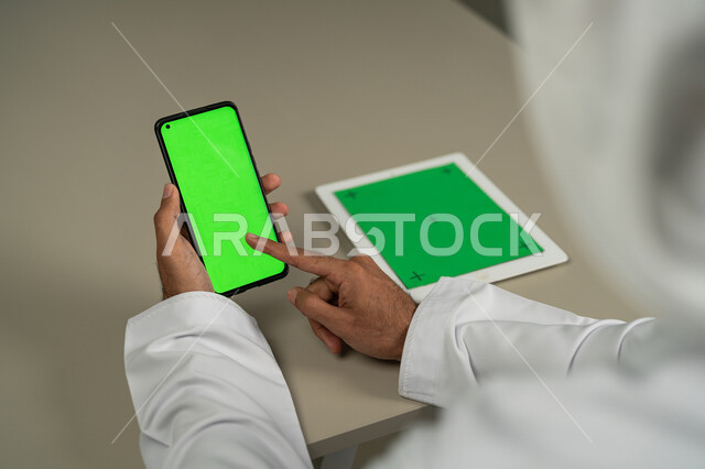 مشاهدة أخبار وسائل التواصل الإجتماعي، التطورات التقنية والتكنولوجيا الحديثة، صورة من الخلف لرجل عربي خليجي اماراتي يستخدم الجوال و التابلت بشاشة خضراء فارغة كروما,  الدردشة والمحادثات مع الأصدقاء, التسويق لموقع إلكتروني، أجهزة الكترونية بشاشة خضراء فارغة