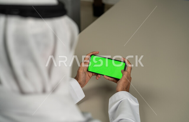 دور التكنولوجيا في في تسهيل الأعمال، عرض شاشة خضراء كروما عبر الهاتف المحمول,  صورة مقربة من الخلف لرجل عربي خليجي اماراتي يستخدم الجوال, الدردشة والمحادثات مع الأصدقاء, التقنيات الحديثة المتطورة, برامج التواصل الاجتماعي, التسويق لموقع إلكتروني