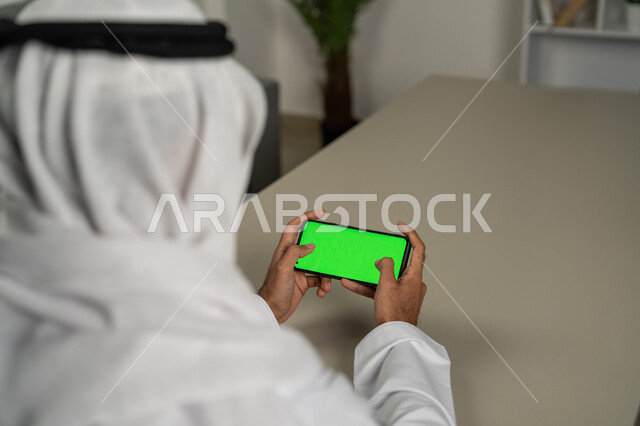 عرض شاشة خضراء كروما عبر الهاتف المحمول,  صورة مقربة من الخلف لرجل عربي خليجي اماراتي يستخدم الجوال, الدردشة والمحادثات مع الأصدقاء, التقنيات الحديثة المتطورة, برامج التواصل الاجتماعي, التسويق لموقع إلكتروني