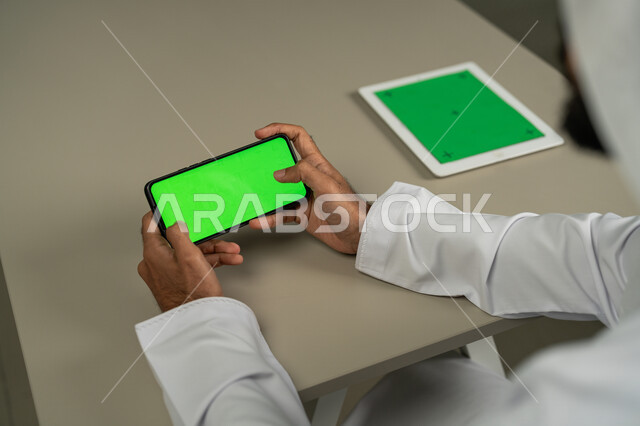 Clicking on a green chroma screen via a mobile phone, a close-up photo from the back of an Arab Gulf Emirati man using a mobile phone, chatting and conversations with friends, advanced modern technologies, social media programs, marketing for a website