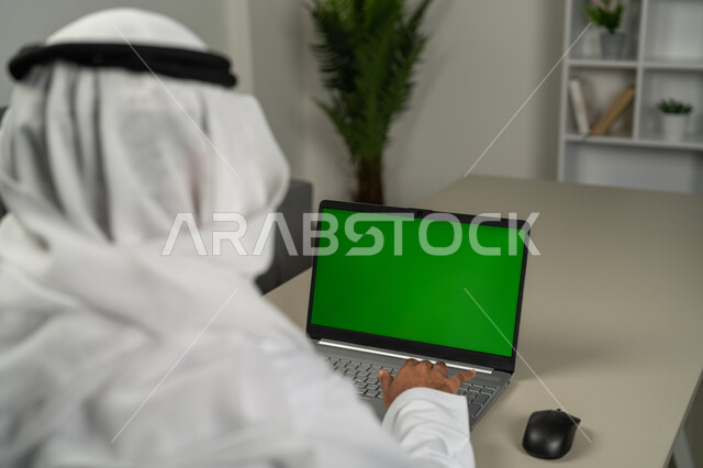 Technical and technological development in various fields, the use of modern devices to complete tasks, displaying a green chroma screen on the laptop, a picture from behind of an Arab Gulf Emirati man wearing a kandora and a white ghutra, chatting and conversations with friends, using technology in remote work.