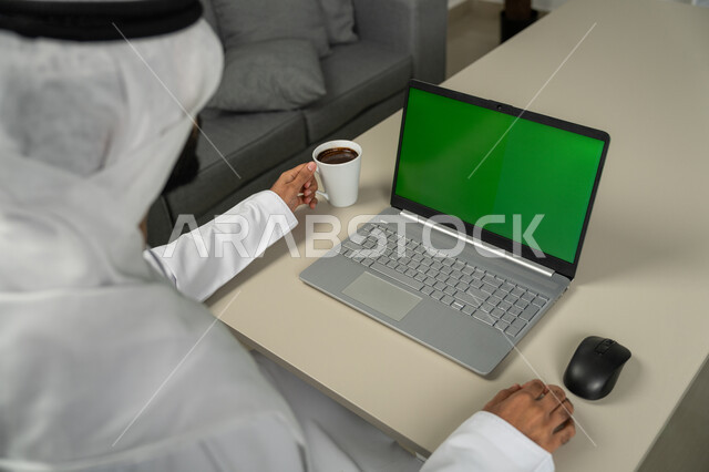 إجراء مقابلات وإجتماعات عن بعد، عرض شاشة خضراء كروما عبر الحاسوب المحمول ، التقنيات الحديثة المتطورة, صورة من الخلف لرجل عربي خليجي اماراتي يرتدي الكندورة والغترة البيضاء يشرب القهوة, برامج التواصل الاجتماعي, الدردشة والمحادثات مع الأصدقاء