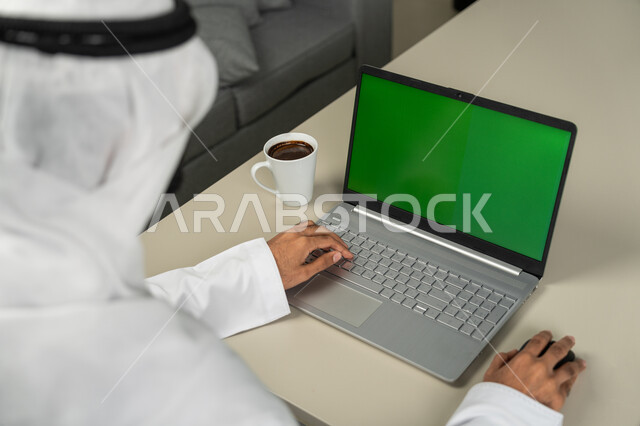 الدردشة والمحادثات مع الأصدقاء، عرض شاشة خضراء كروما عبر الحاسوب المحمول ، التقنيات الحديثة المتطورة, صورة من الخلف لرجل عربي خليجي اماراتي يرتدي الكندورة والغترة البيضاء يشرب القهوة, برامج التواصل الاجتماعي, إجراء مقابلات وإجتماعات عن بعد