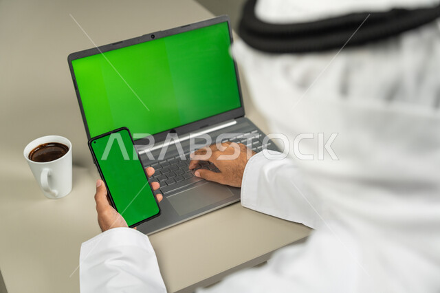 عرض شاشة خضراء كروما عبر الحاسوب المحمول والهاتف، التقنيات الحديثة المتطورة, صورة من الخلف لرجل عربي خليجي اماراتي يرتدي الكندورة والغترة  يمسك الموبايل بيده ويشرب القهوة, الدردشة والمحادثات مع الأصدقاء, برامج التواصل الاجتماعي, إجراء مقابلات وإجتماعات عن بعد