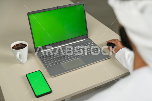 التقنيات الحديثة المتطورة, عرض شاشة خضراء كروما عبر الحاسوب المحمول و الهاتف ,صورة من الخلف لرجل عربي خليجي اماراتي يرتدي الكندورة و الغترة البيضاء يشرب القهوة, الدردشة والمحادثات مع الأصدقاء, برامج التواصل الاجتماعي, إجراء مقابلات وإجتماعات العمل عن بعد