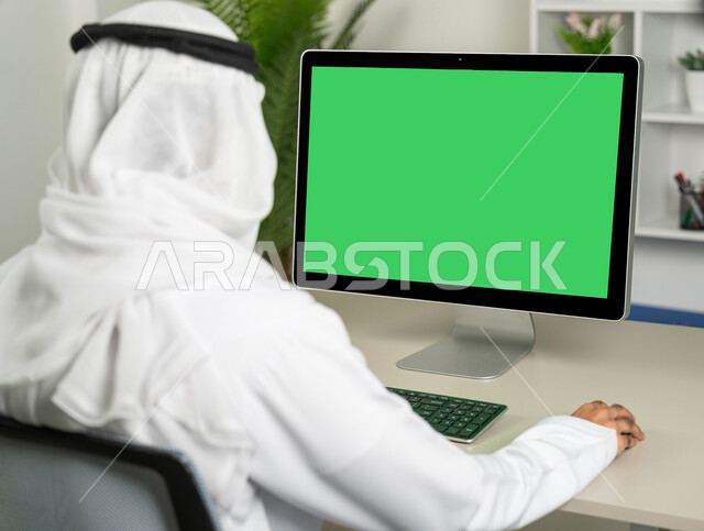 Using a desktop computer with a green chroma background, chatting, conversations, and social networking, marketing a website, a picture from the back of an Arab Gulf Emirati man wearing a kandora and a white ghutra, drinking coffee, making a video call, watching movies and series.