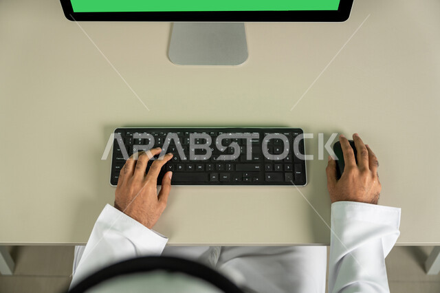 Making a video call, using a desktop computer with a green chroma background, chatting, conversations, and social networking, marketing a website, a picture from above of an Arab Gulf Emirati man wearing a kandora and a white ghutra, watching movies and series.
