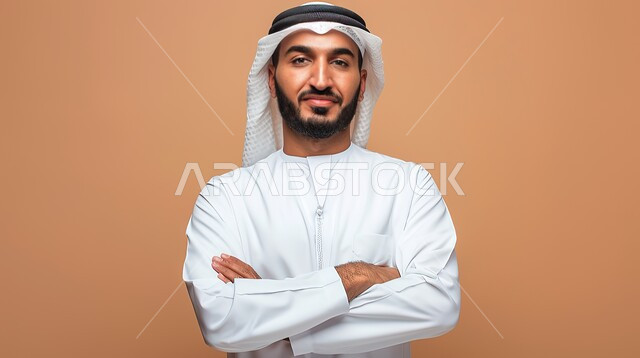 مفهوم الاناقة والرجولة، الاهتمام بالمظهر الخارجي، النظر إلى الكاميرا بايماءات الثقة بالنفس، بورتريه مقرب لرجل عربي خليجي اماراتي يرتدي الزي التقليدي يقف بايدي متقاطعة، خلفية بنية