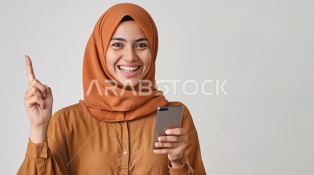 استخدام الاجهزة التكنولوجية الحديثة، سماع الاخبار والمفاجأت السعيدة، ايماءات الدهشة والسرور، بورتريه لإمرأة عربية خليجية سعودية محجبة مبتسمة تنظر إلى الكاميرا وتحمل هاتفها المحمول بتعابير الفرح والسعادة، الدردشة والتواصل مع الاهل والاصدقاء، خلفية رمادية
